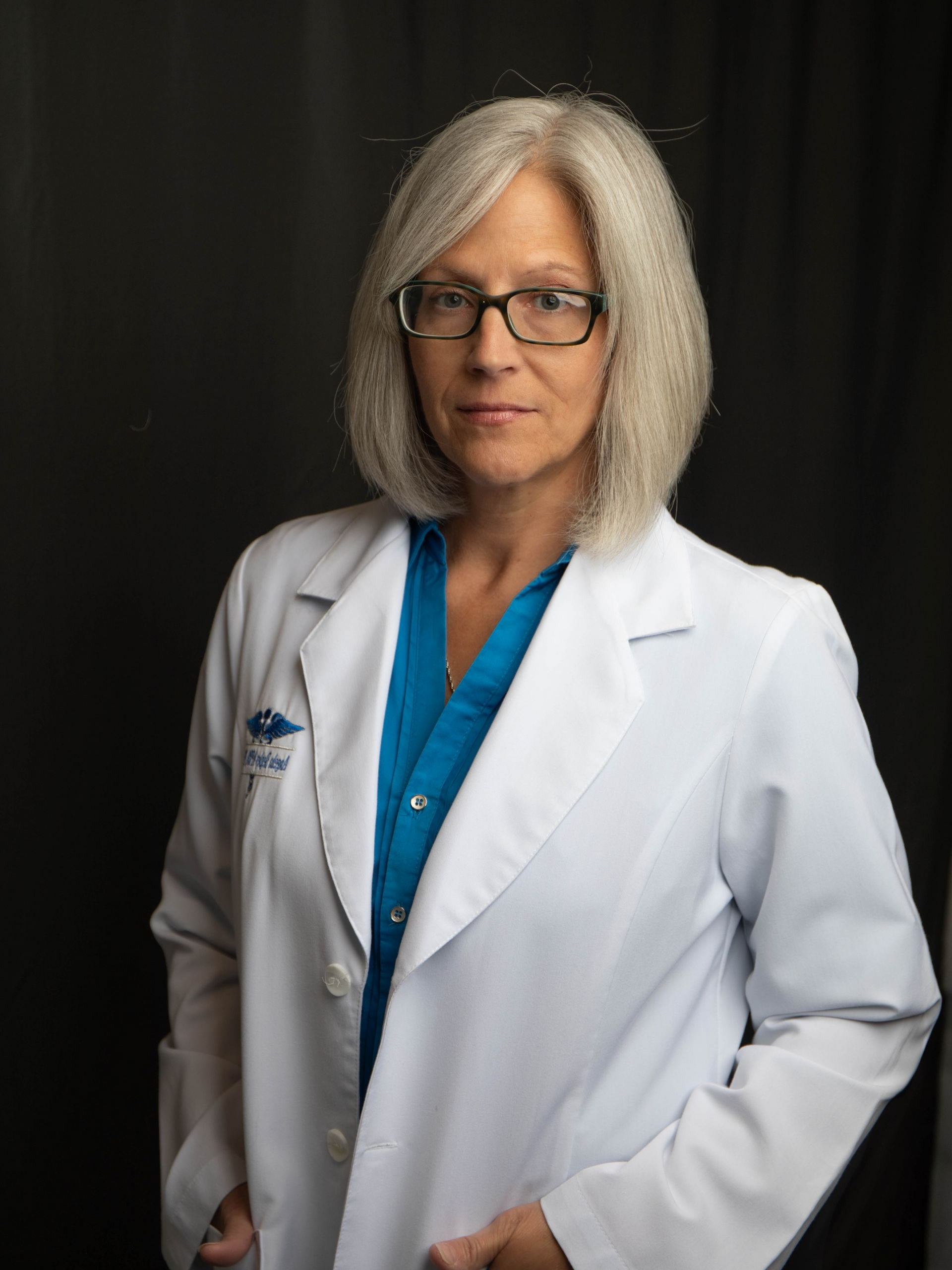 Woman in a white lab coat and glasses standing against a dark background.