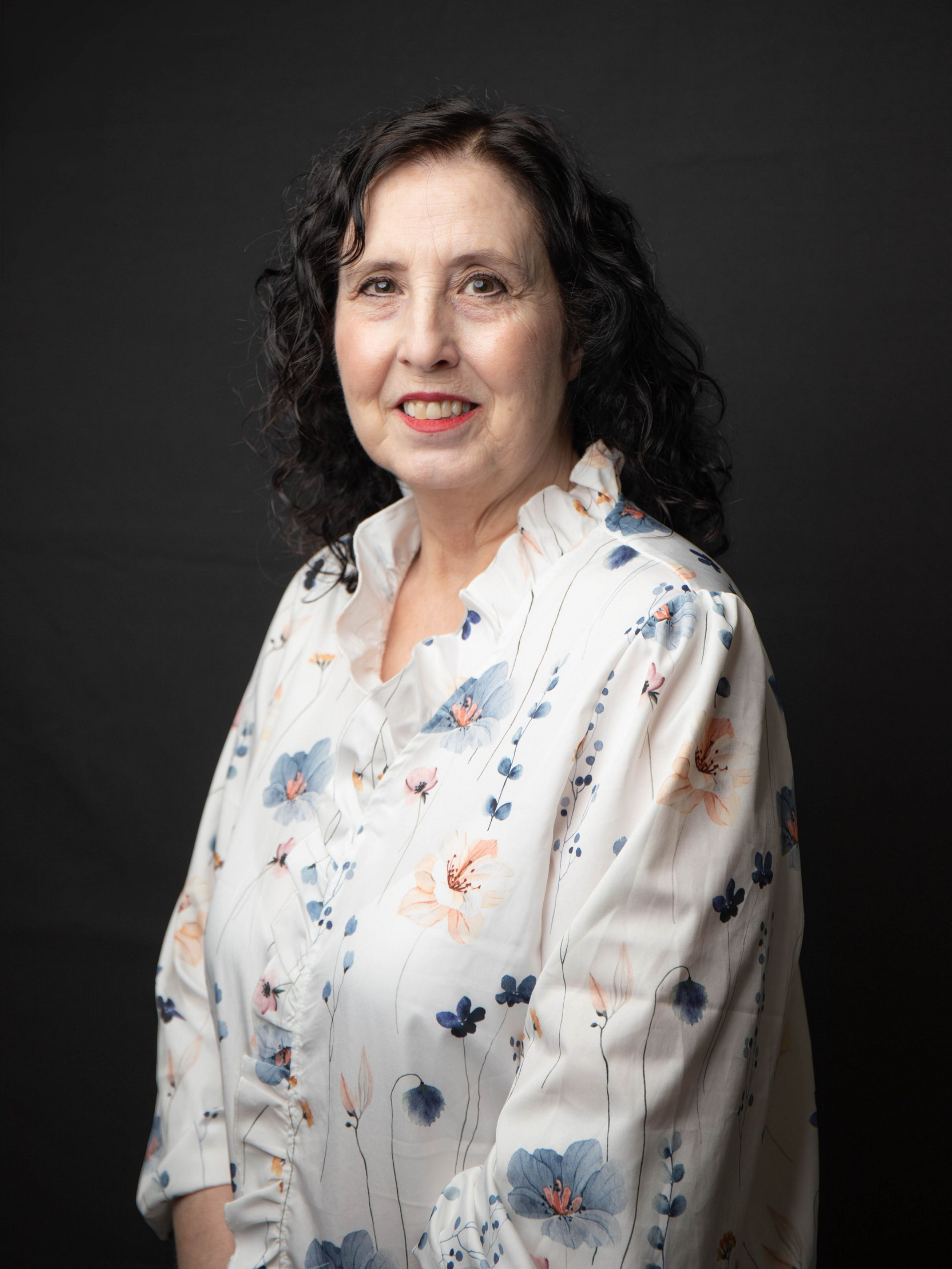 Portrait of a woman with dark curly hair wearing a black and blue floral top against a dark background