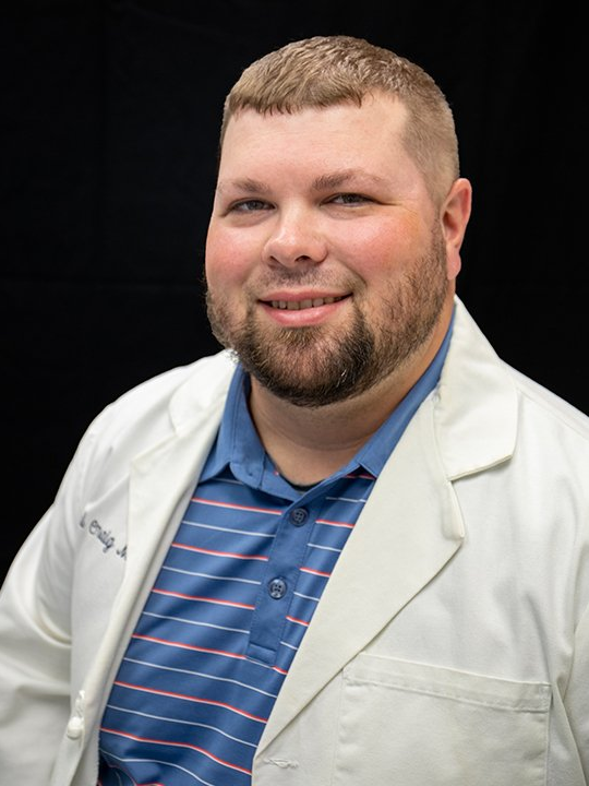 Smiling man in a white lab coat over a blue striped polo against a black background