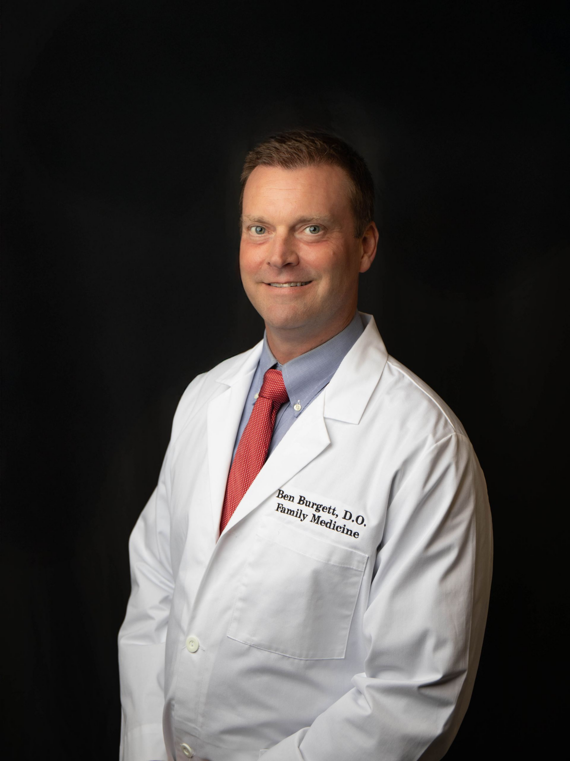 Doctor in white coat with teal stethoscope and yellow tie, standing against a black background