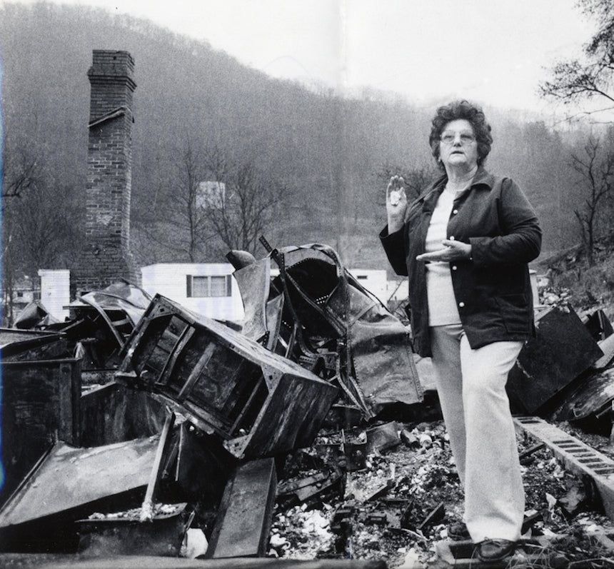Woman stands amid burned wreckage in front of a damaged chimney and hillside.