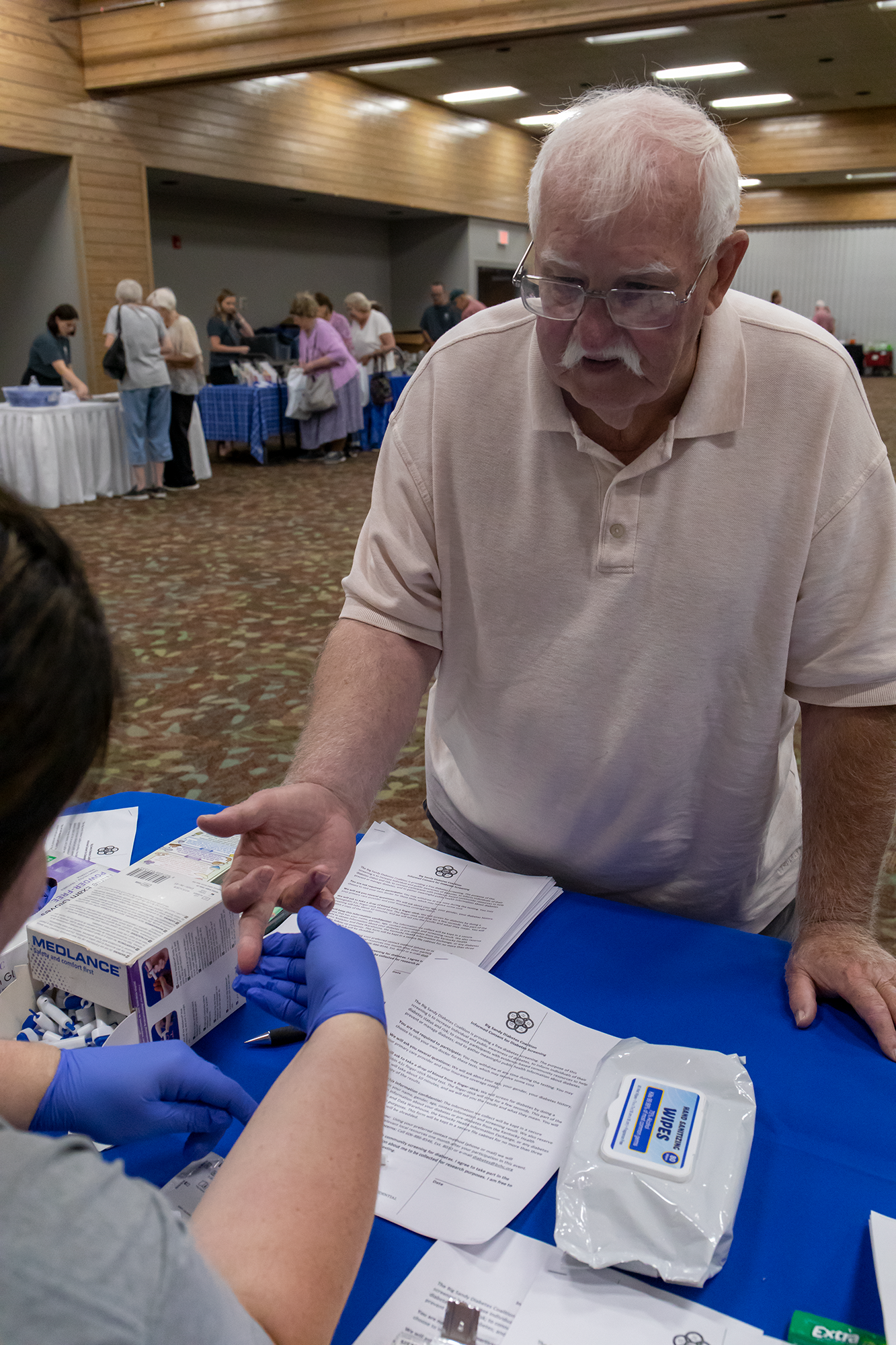 Man in beige polo speaking to a person in blue gloves at a table with packaged items in a community hall