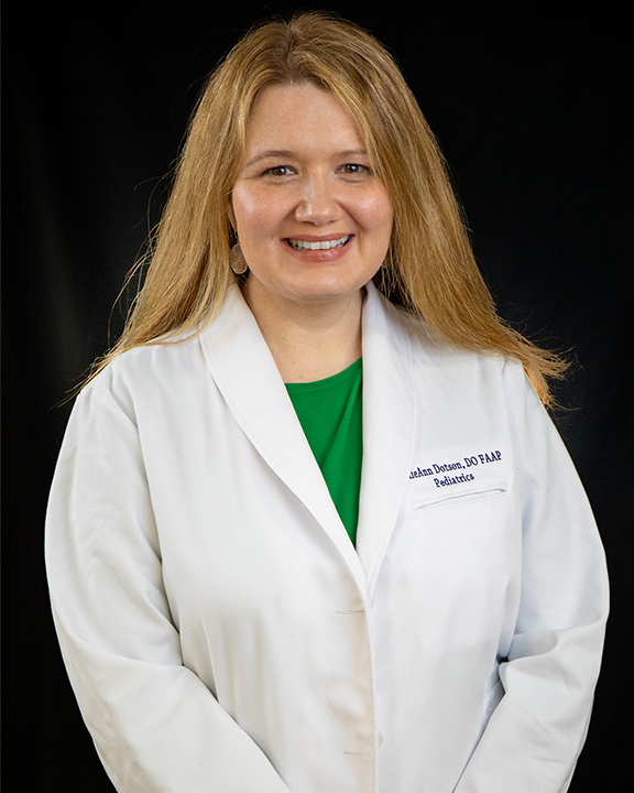 Woman in a white lab coat over a green shirt, smiling against a black background.