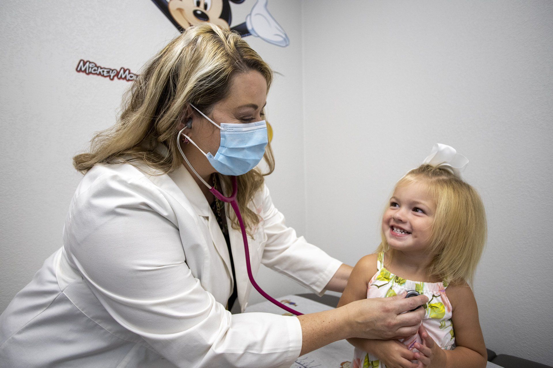 Doctor in white coat and mask examining a smiling child in a clinic room