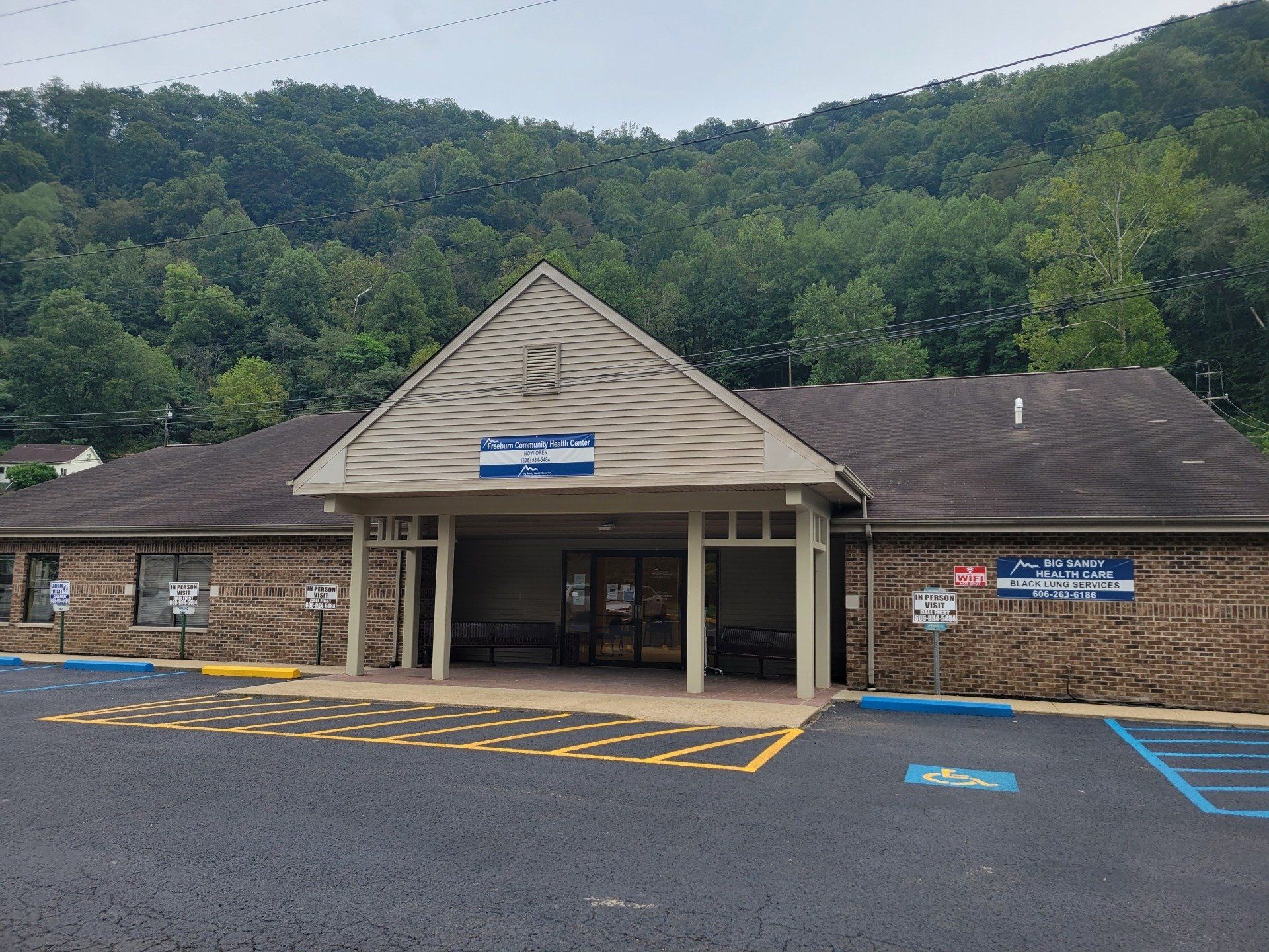 Brick building entrance with covered drive-through, blue parking spaces, and wooded hillside behind.