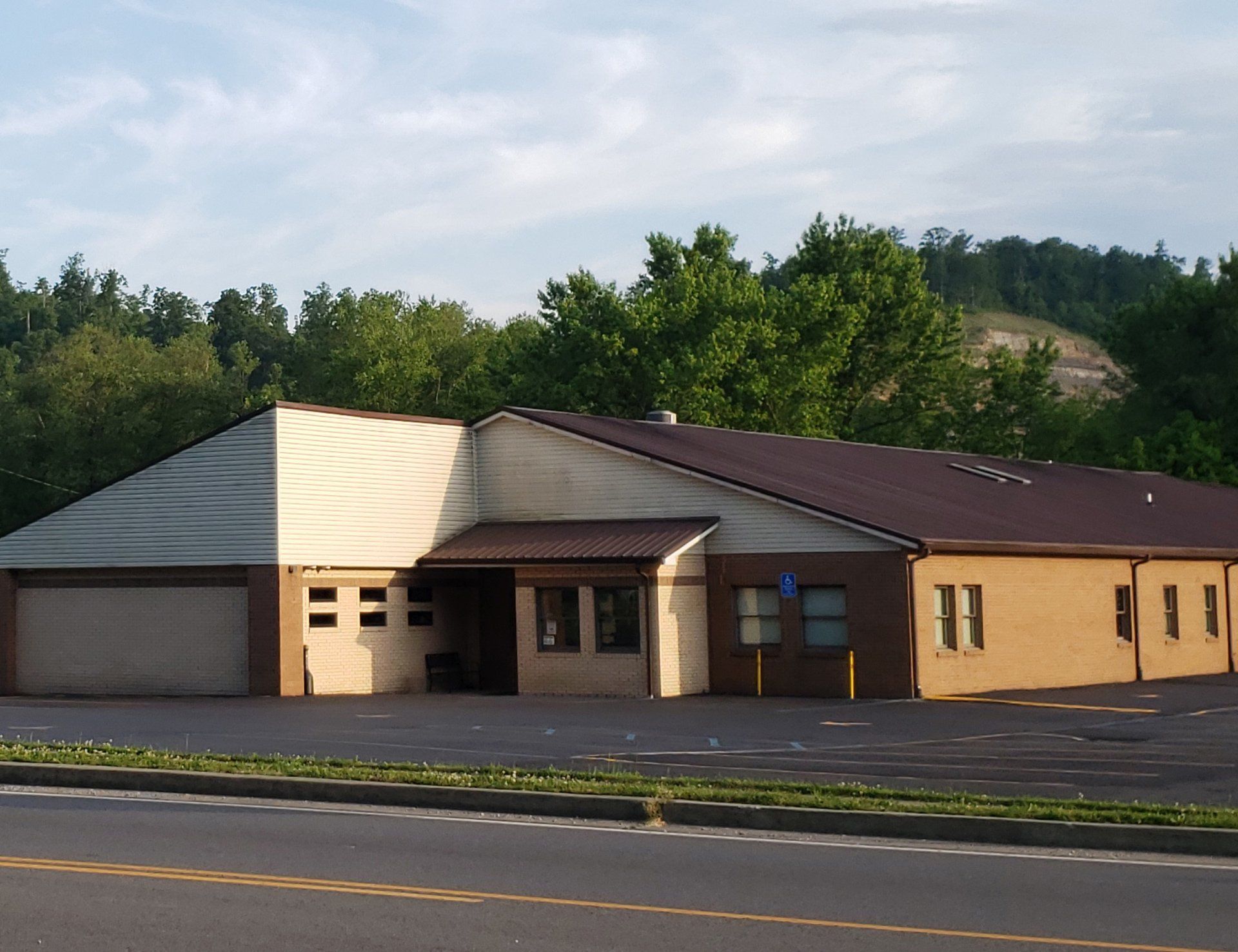 Single-story tan building with a brown roof beside a road, surrounded by trees and hills.
