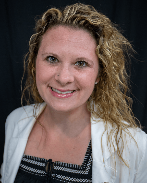 Smiling woman with curly blonde hair, wearing a white blazer and patterned top against a dark background