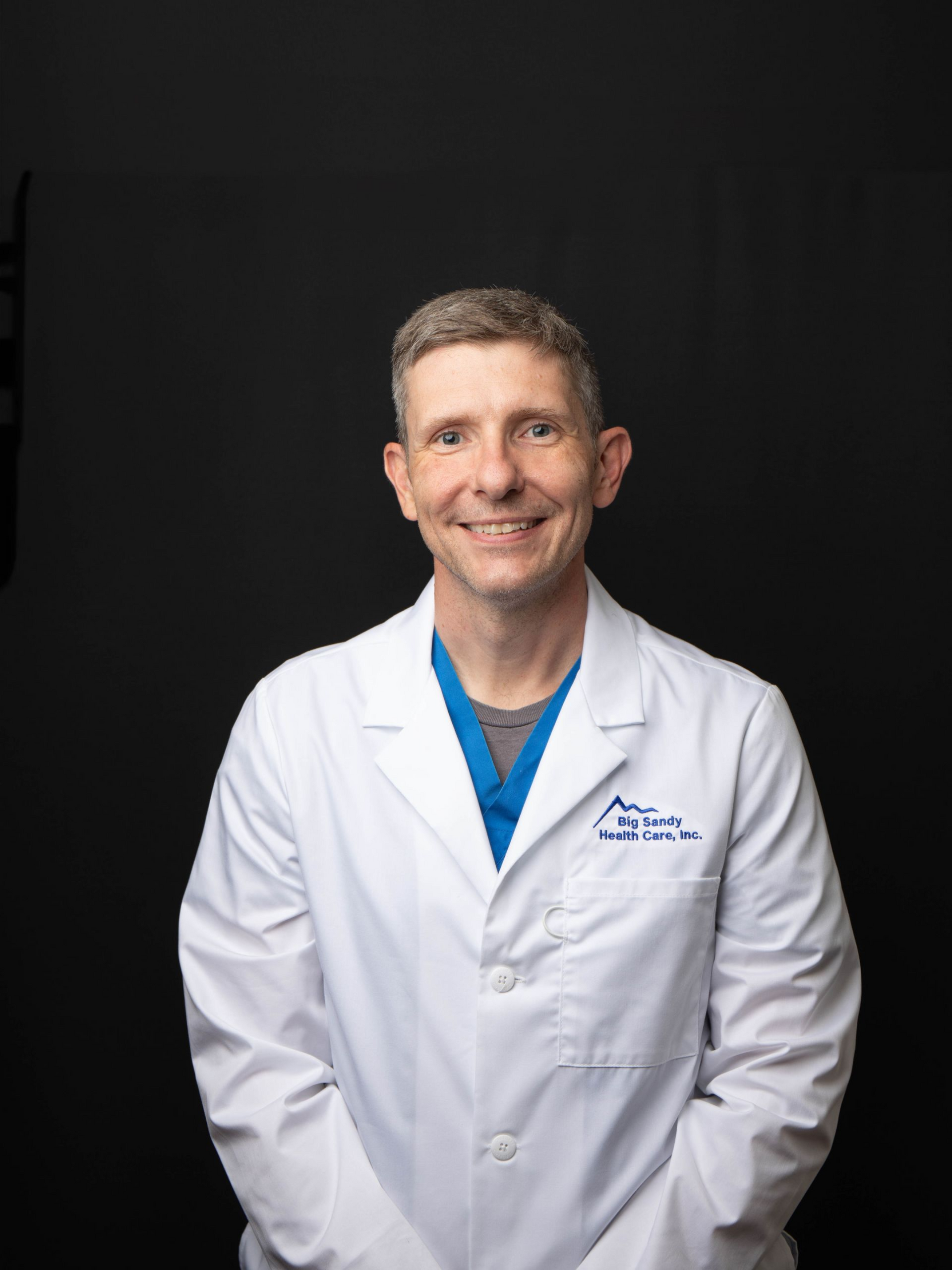 Smiling medical professional in a white coat and teal scrubs against a dark background
