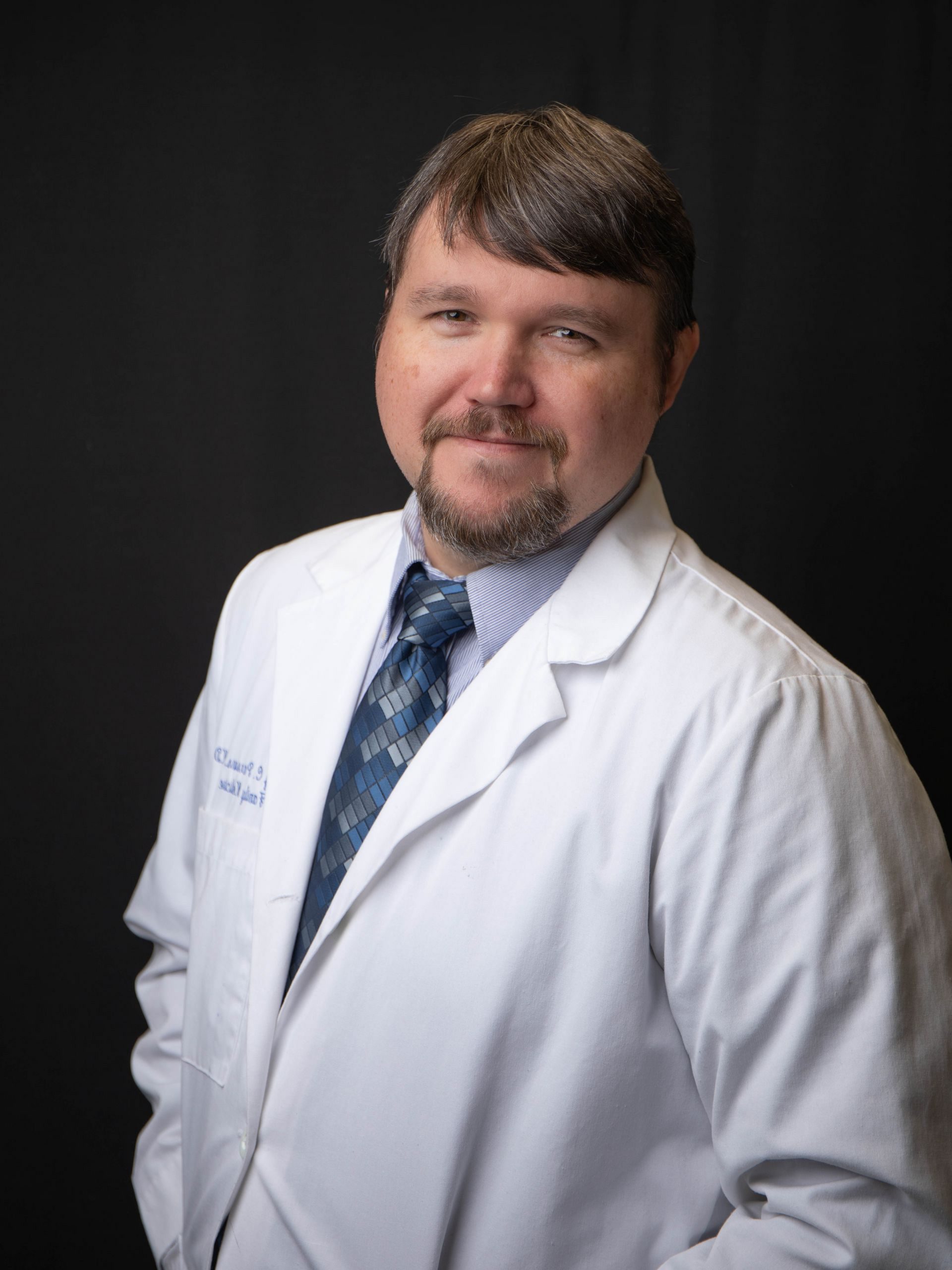 Smiling doctor in white coat and tie, posed against a black background.