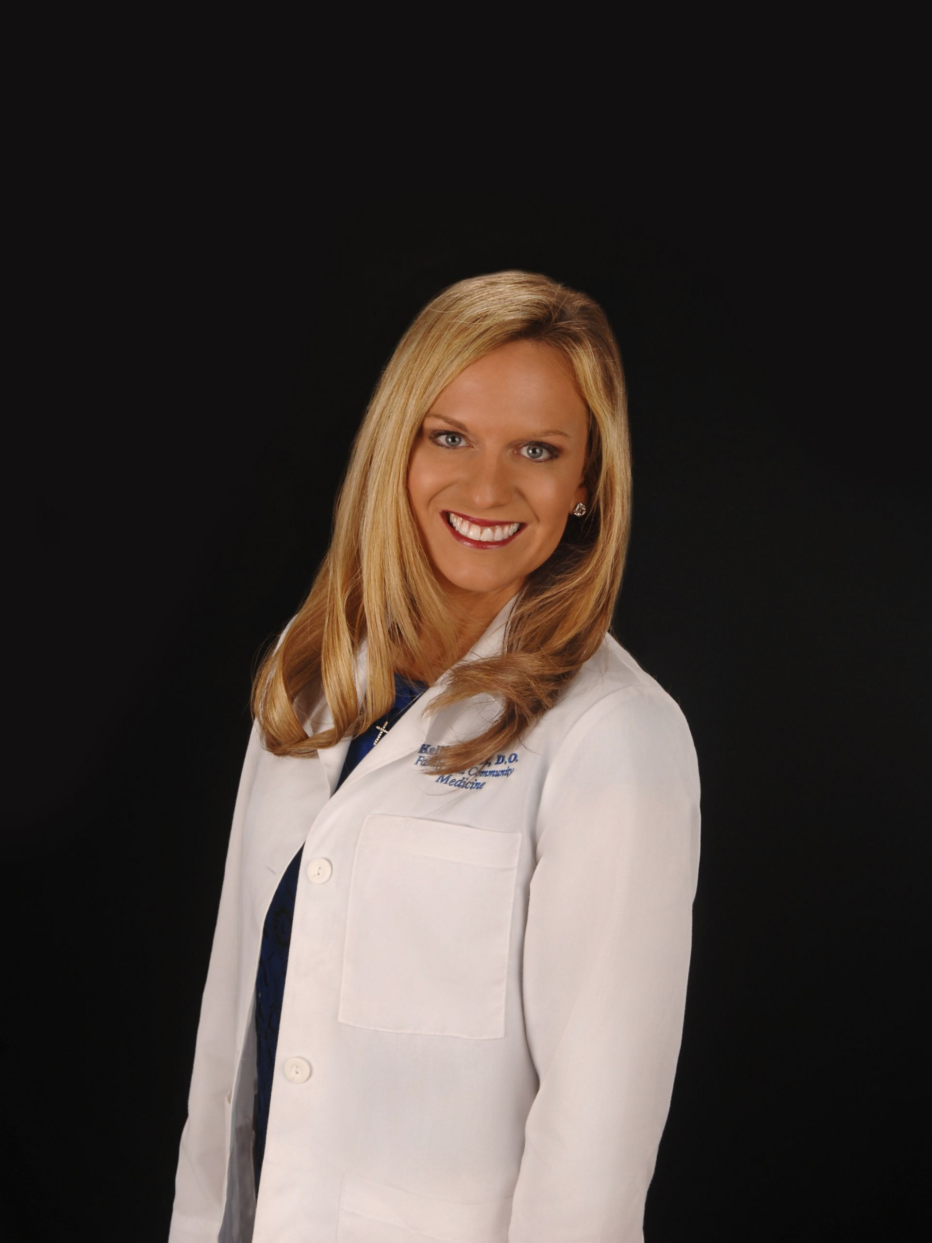 Doctor in a white coat with stethoscope and pink shirt, smiling against a dark background.