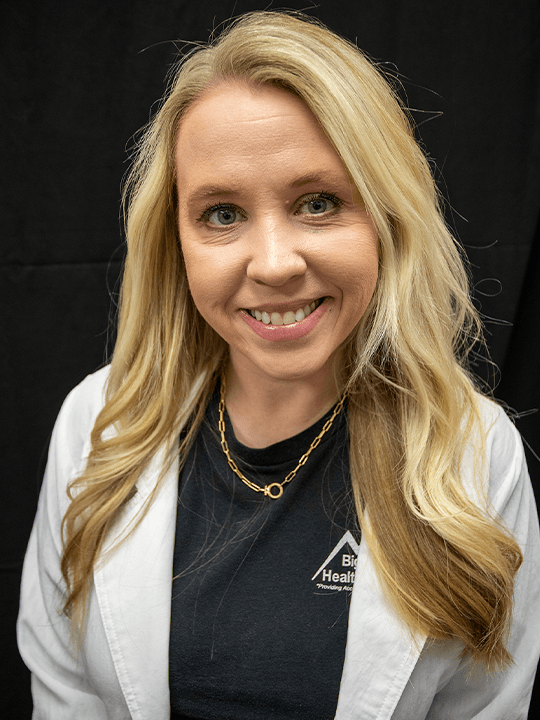 Smiling blonde woman in a white lab coat over a dark shirt against a black background