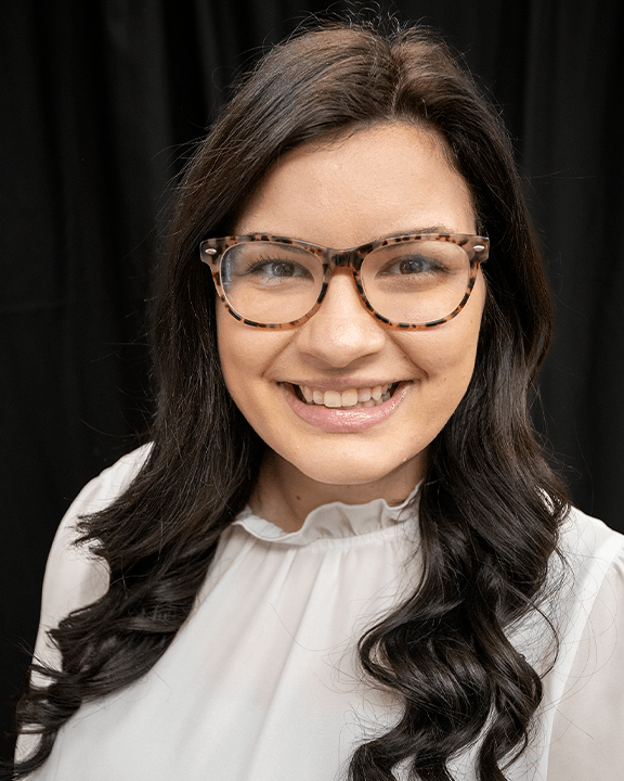 Smiling person with long dark hair and glasses, wearing a white blouse against a black background.