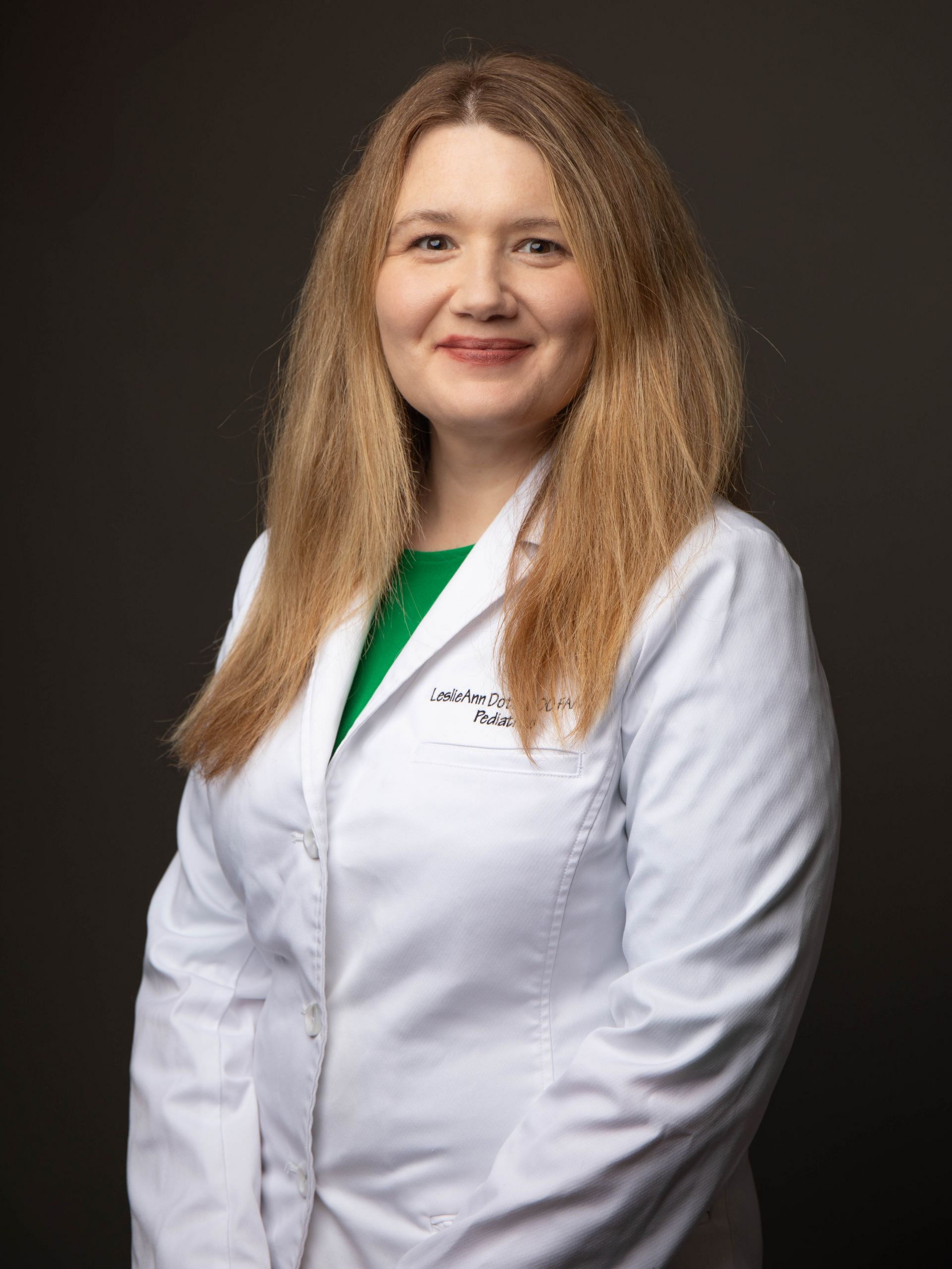 Woman in a white lab coat over a green shirt, smiling against a black background.