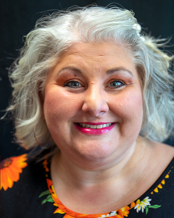 Smiling woman with silver hair in a black floral top against a dark background
