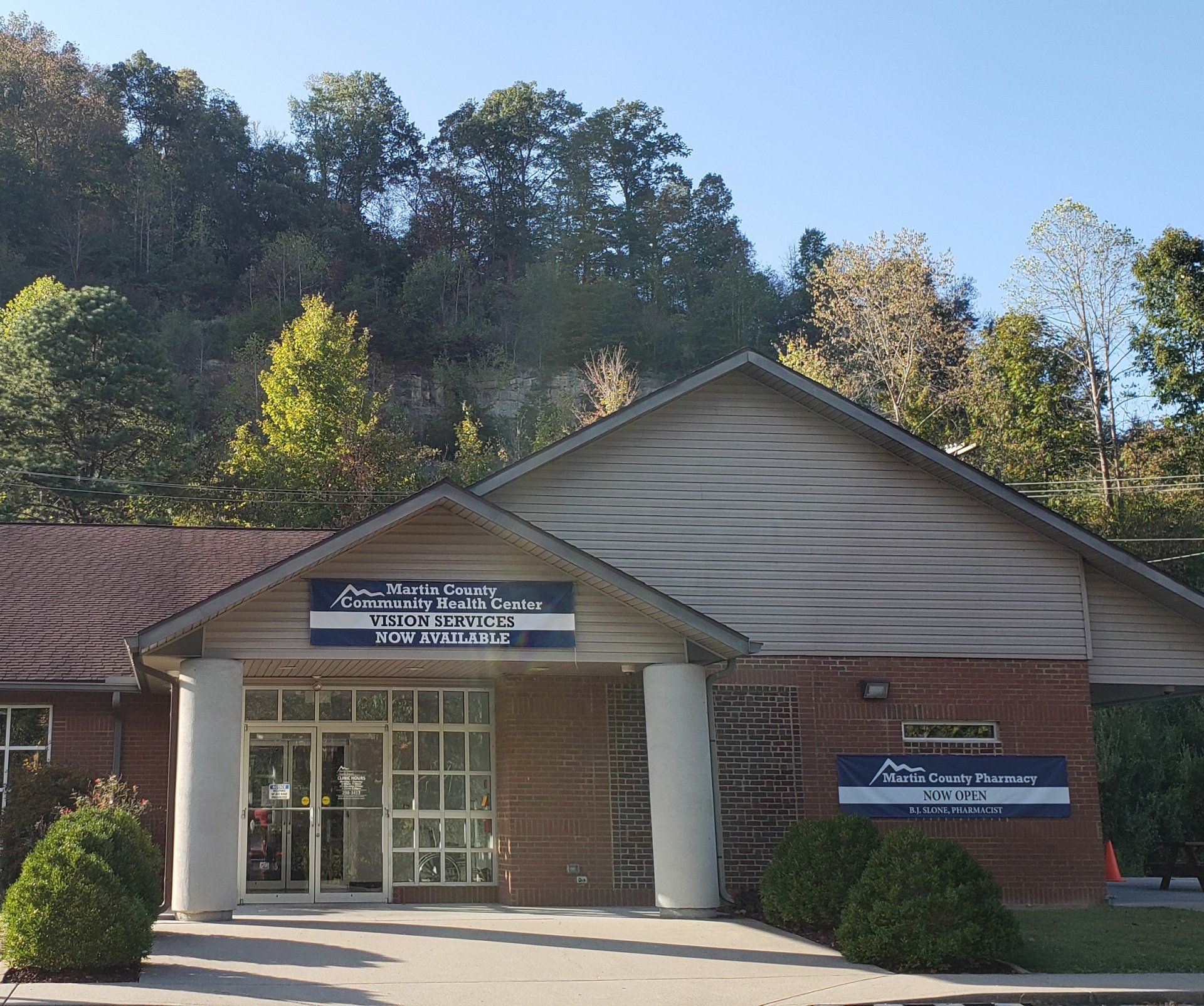 Brick-and-white entrance building with blue signs, set against a wooded hillside under a clear sky