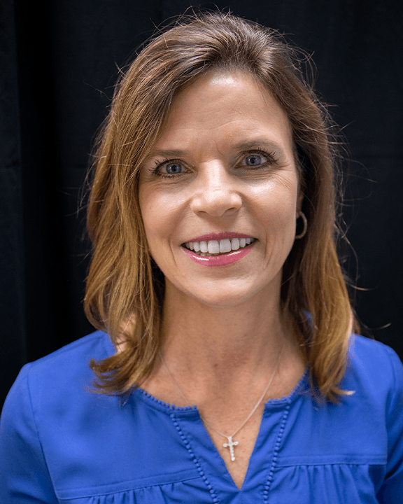 Woman in a blue top smiling against a dark background.