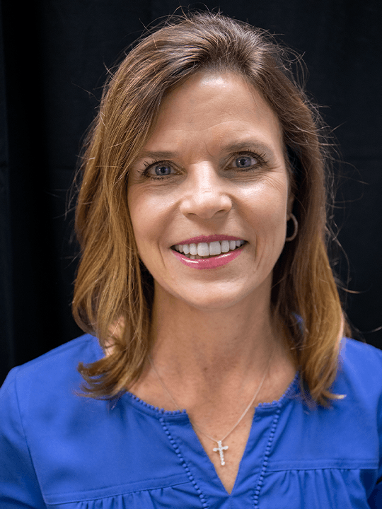 Smiling woman in a blue blouse with a cross necklace against a dark background