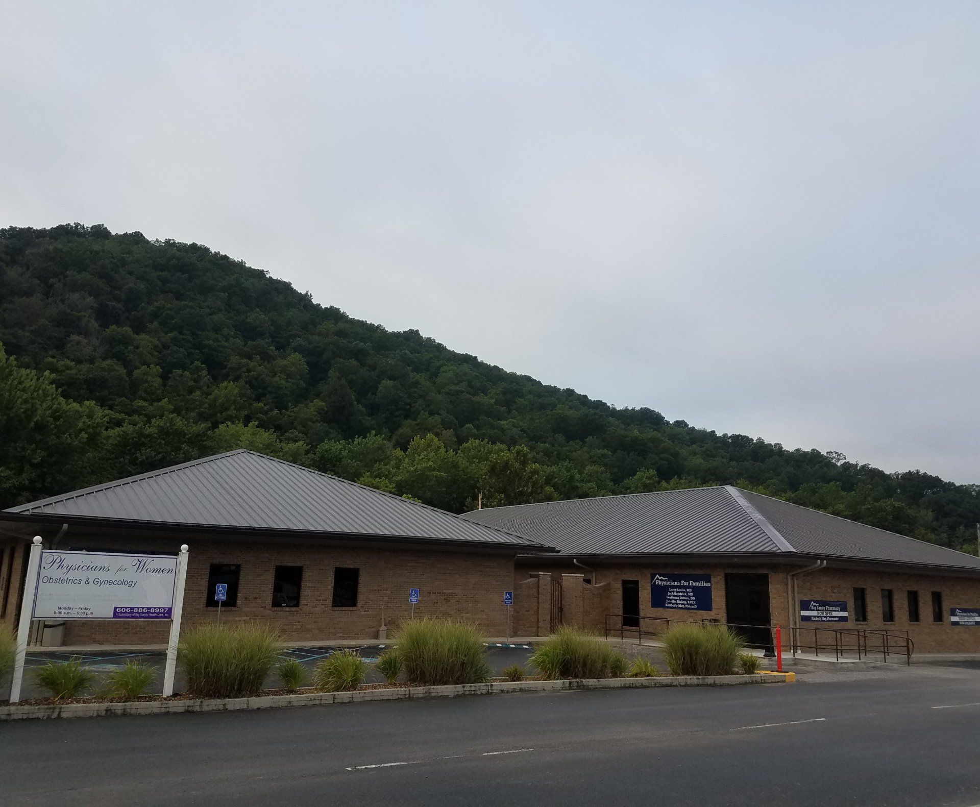 Single-story brick building with gray roof beside a forested hillside and parking lot under cloudy skies