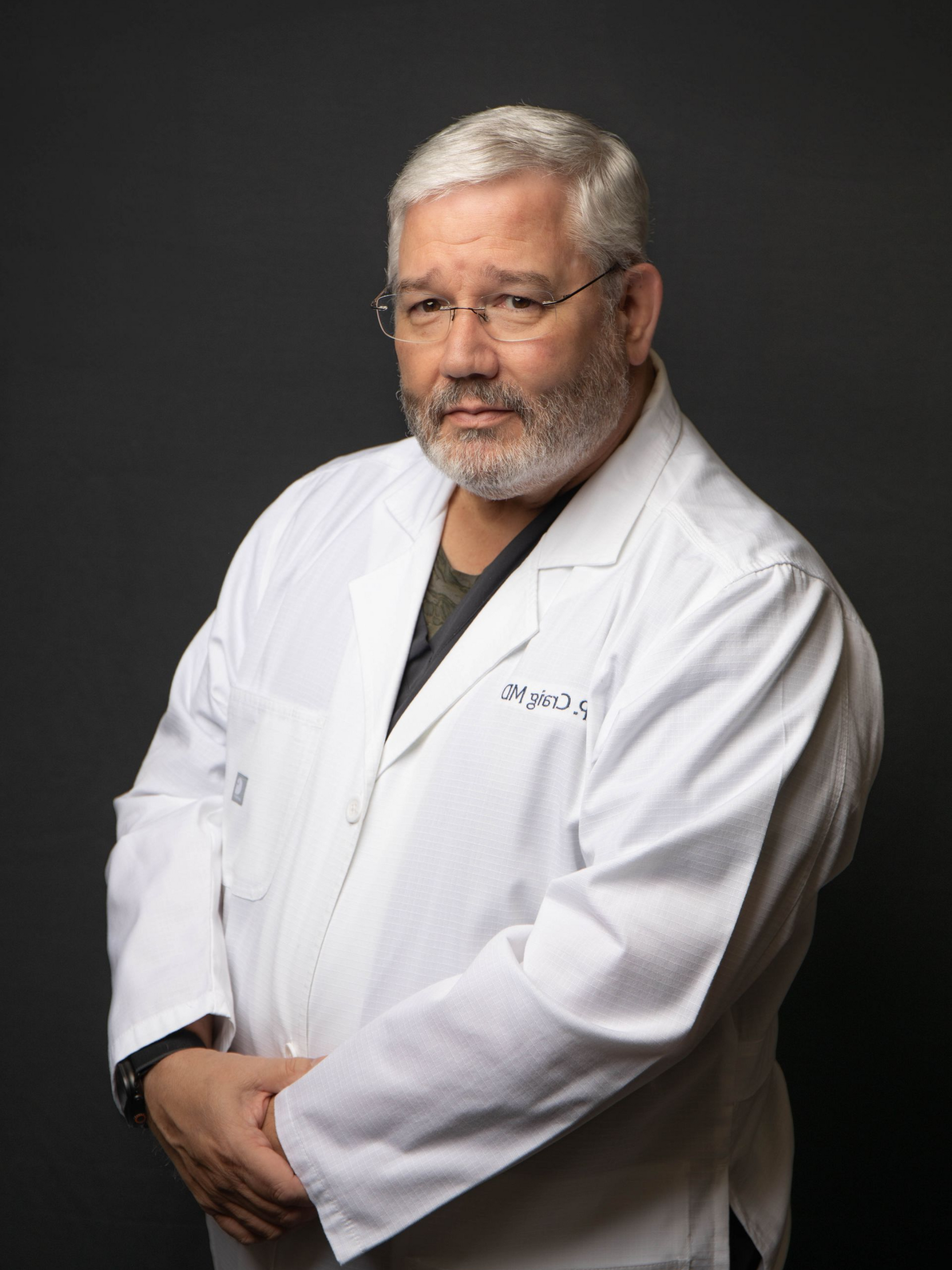 Doctor in a white lab coat with a name badge, standing against a dark background, looking at the camera.