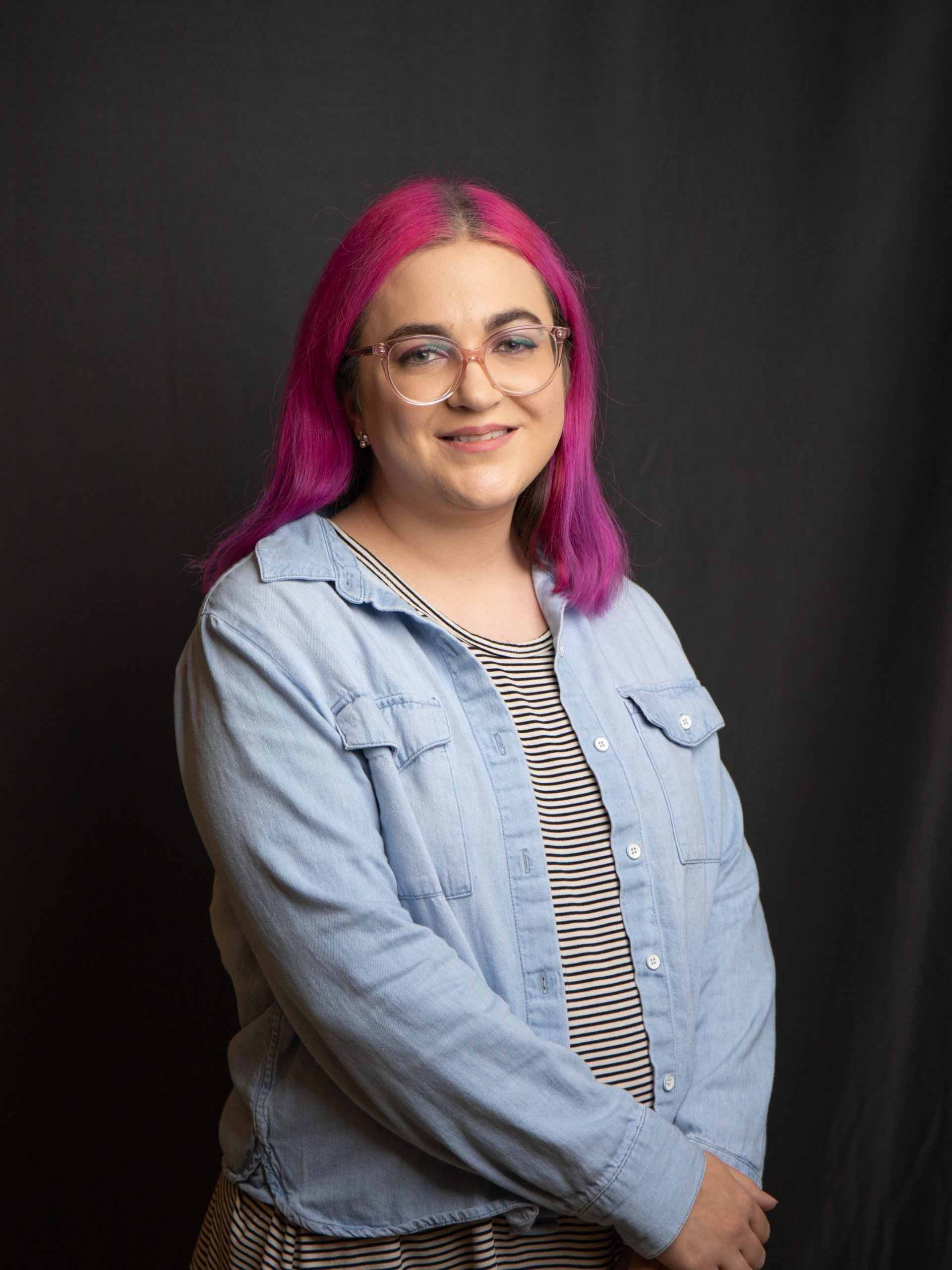 Portrait of a smiling person with blue hair, glasses, and a striped shirt against a dark background