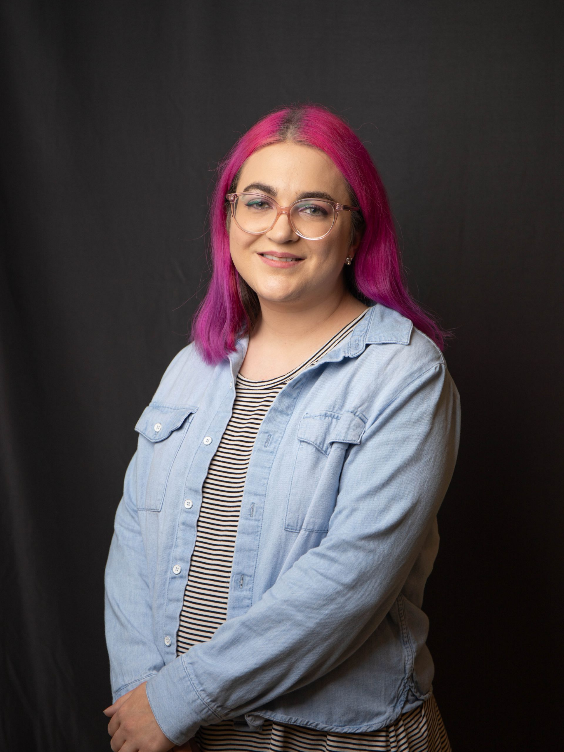 Portrait of a smiling person with blue hair, glasses, and a striped shirt against a dark background