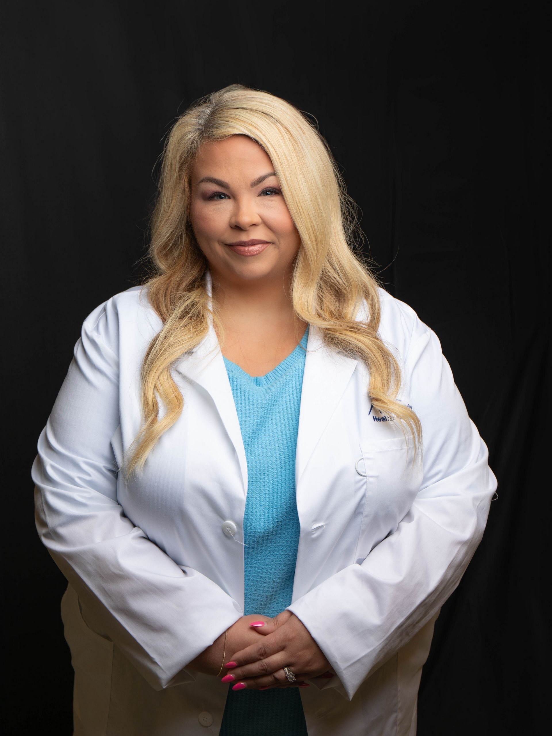 Blonde doctor in white coat and blue blouse, smiling against a dark background.