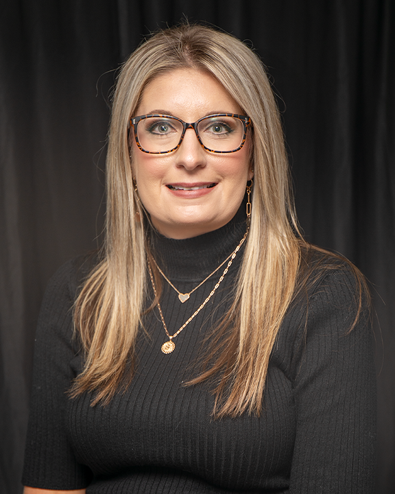 Portrait of a smiling woman with glasses in a black top against a dark backdrop