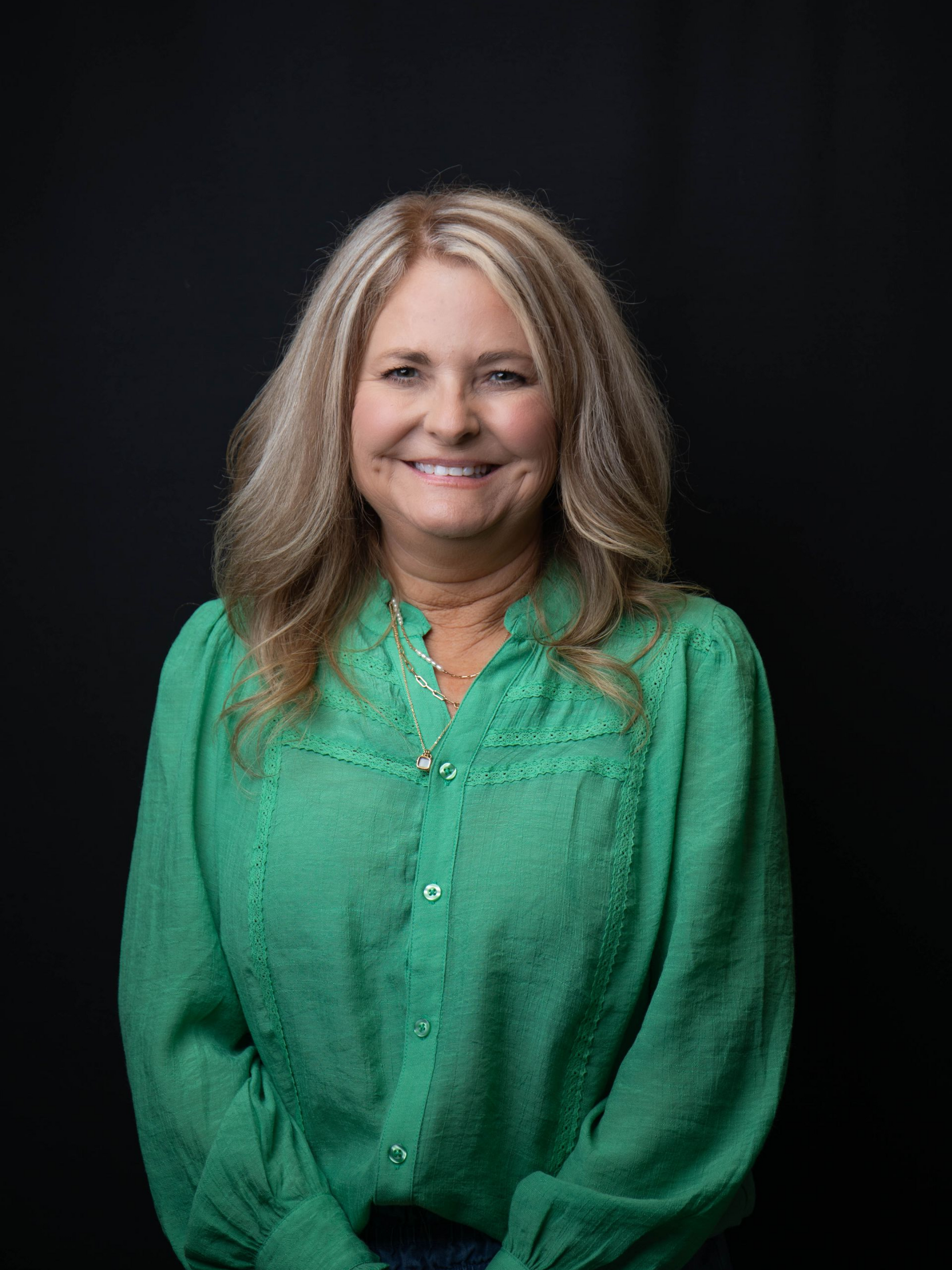 Smiling woman in a white coat and turquoise top, wearing pearl necklaces, against a dark background