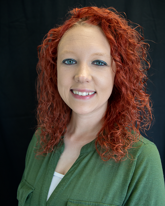 Smiling person with curly red hair wearing a green top against a dark background