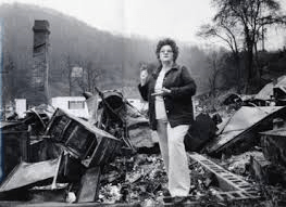 Person standing amid fire-damaged debris in a snowy outdoor scene with a smoke-stained chimney.