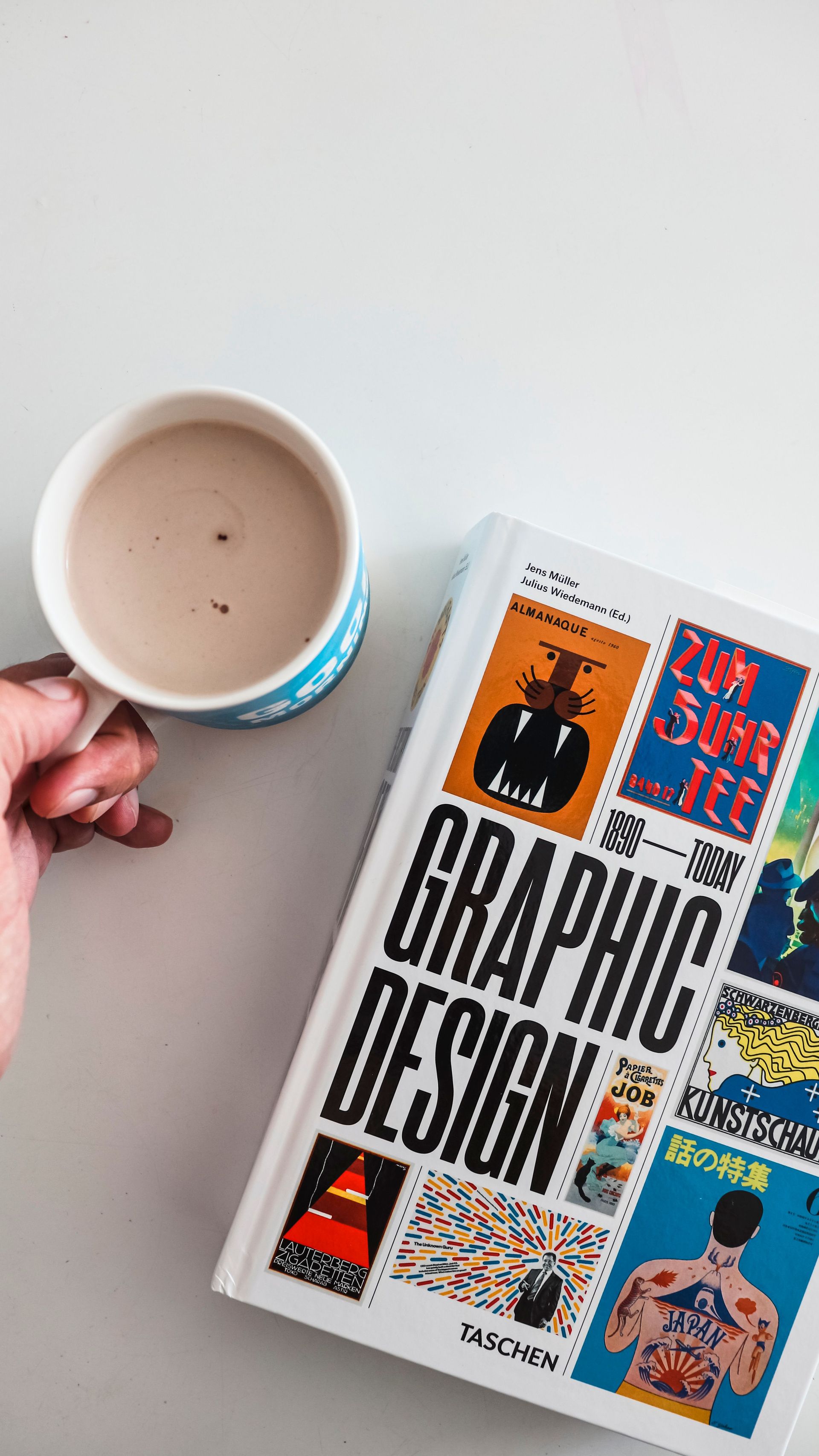 Hand holding a coffee mug beside a book titled “Graphic Design” on a white table.