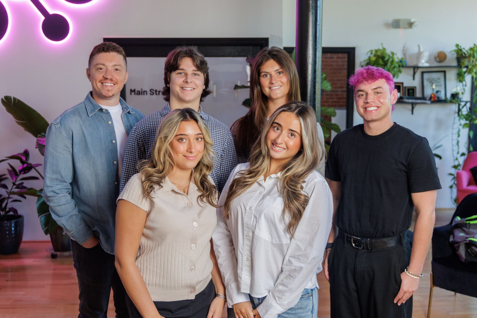 Six people posing indoors in front of a purple-lit wall and plants, smiling at the camera