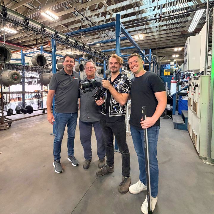 Four people posing in an industrial workshop, smiling beside machinery and steel beams.