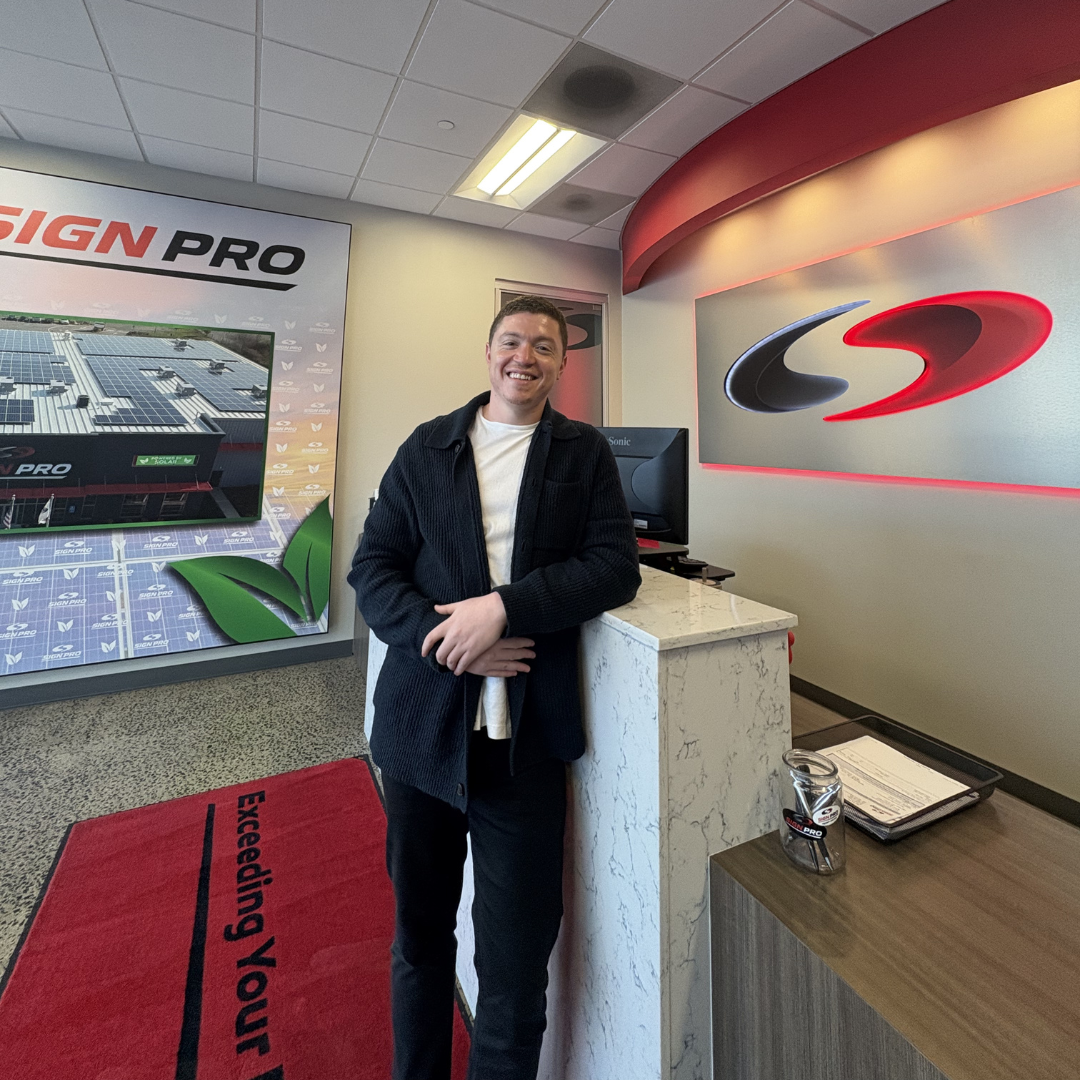 Man in a black jacket smiles at the front desk of an office, which has a logo with red and silver details.