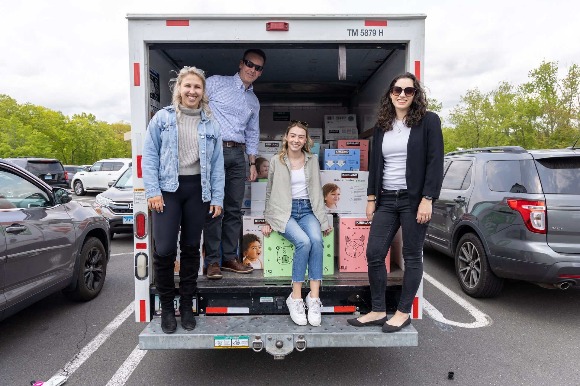 Four people stand at the back of a delivery truck in a parking lot, smiling. Boxes are inside.