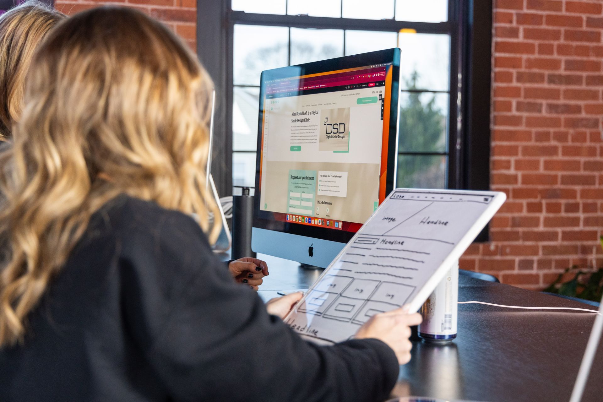 Person holding a clipboard at a desk, looking at a desktop monitor in a brick-walled office