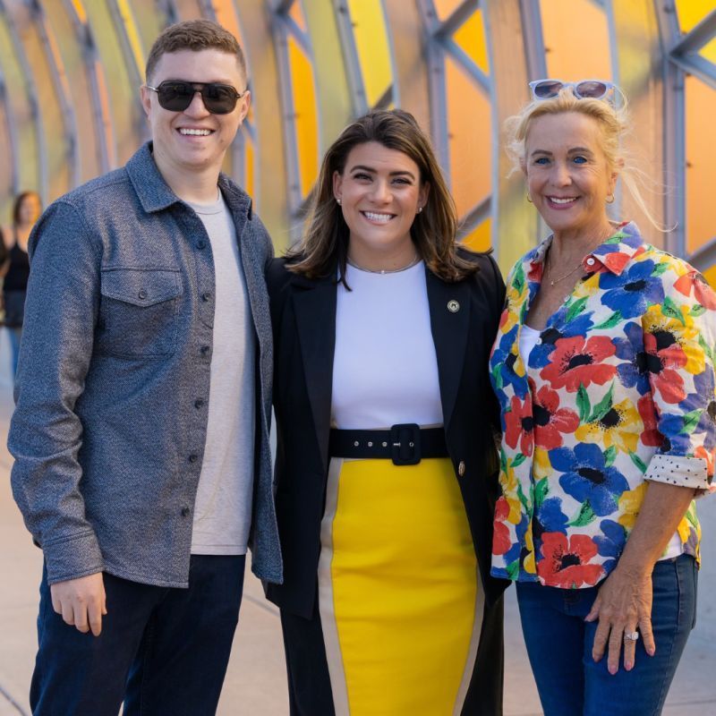 Three people smiling outdoors: woman in blazer, woman in floral shirt, and man in denim shirt. Colorful background.
