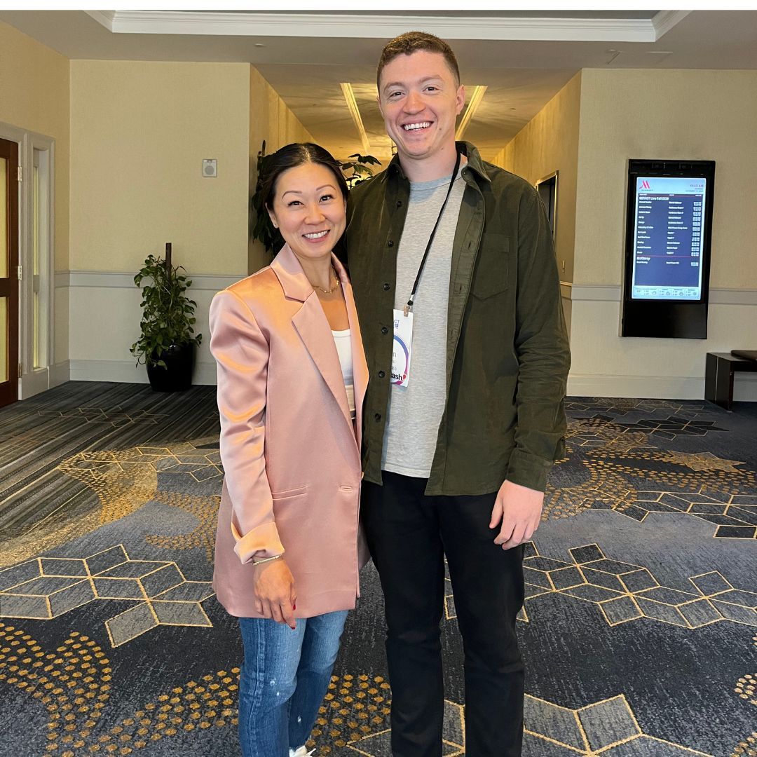 A woman and a man standing in a hallway. The woman wears a pink blazer and jeans. The man wears a green jacket and black pants.