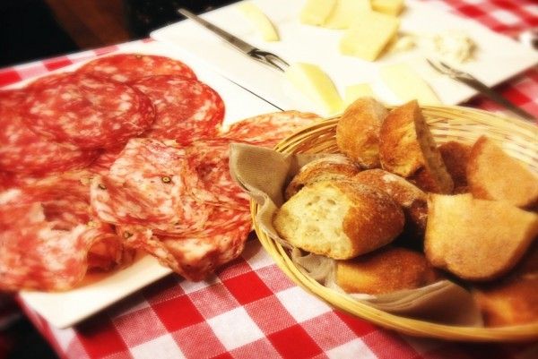 Fette di salame e grissini su una tovaglia a quadretti, con del formaggio sullo sfondo