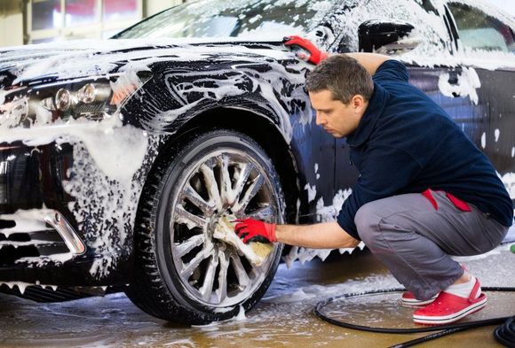 Man washes a black car's wheel with a sponge, covered in soap suds, outdoors, wearing red gloves and shoes.