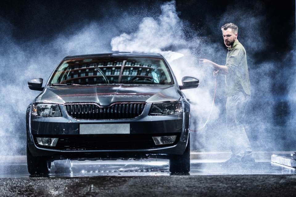 Man washing a dark gray car in a mist-filled setting.