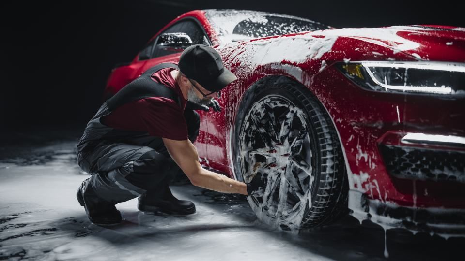 Person washing a red car covered in soapy foam, scrubbing a wheel in a dark setting.