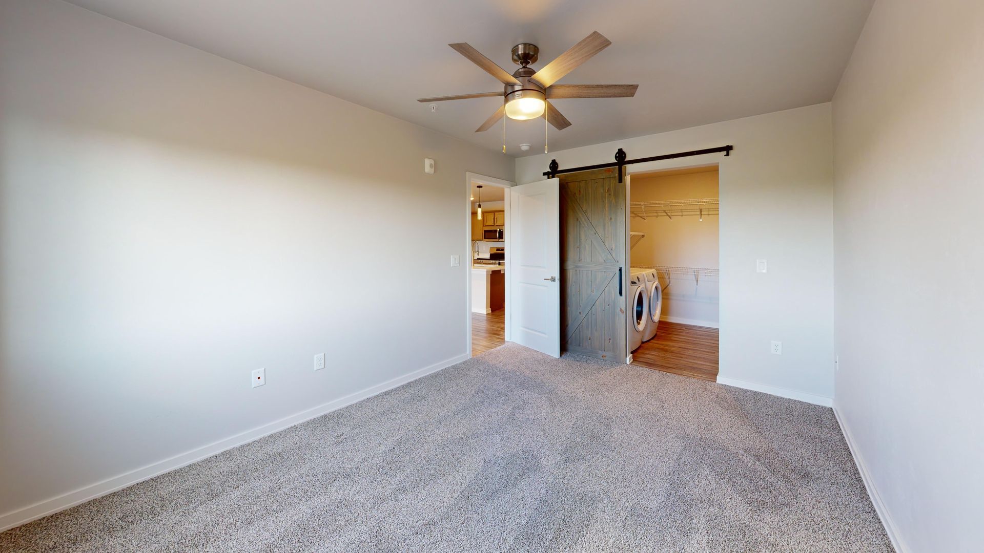 Empty beige bedroom with gray carpet, ceiling fan, and barn-door closet opening to a bathroom.