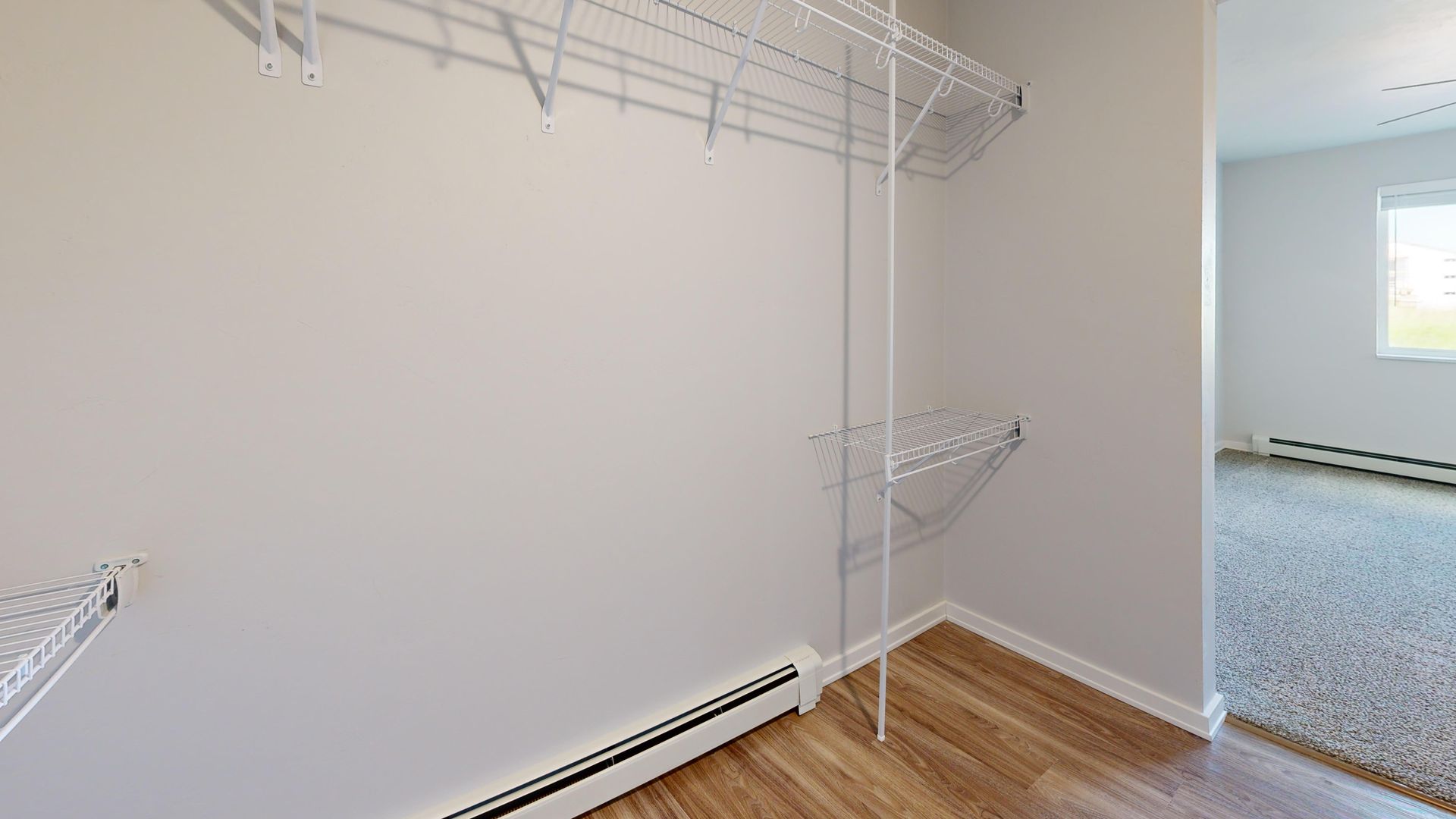 Empty white closet with wire shelves, hardwood floor, and doorway to a carpeted room