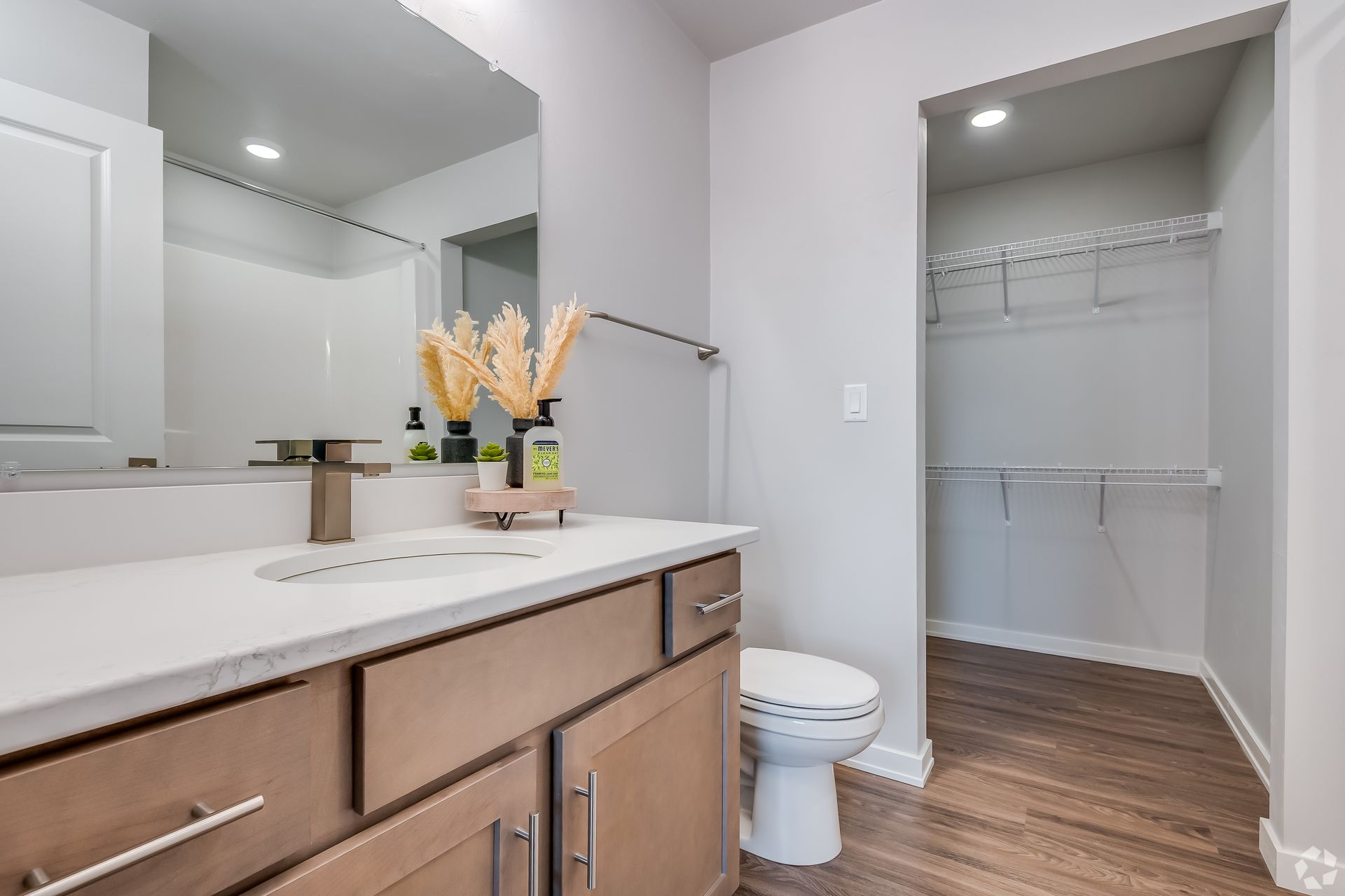Bright bathroom with wood vanity, large mirror, toilet, and open closet with shelving.
