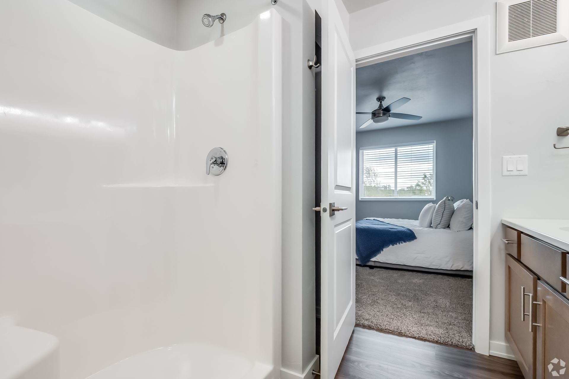 Bathroom view into a bedroom with white walls, a ceiling fan, and a bed with blue bedding.