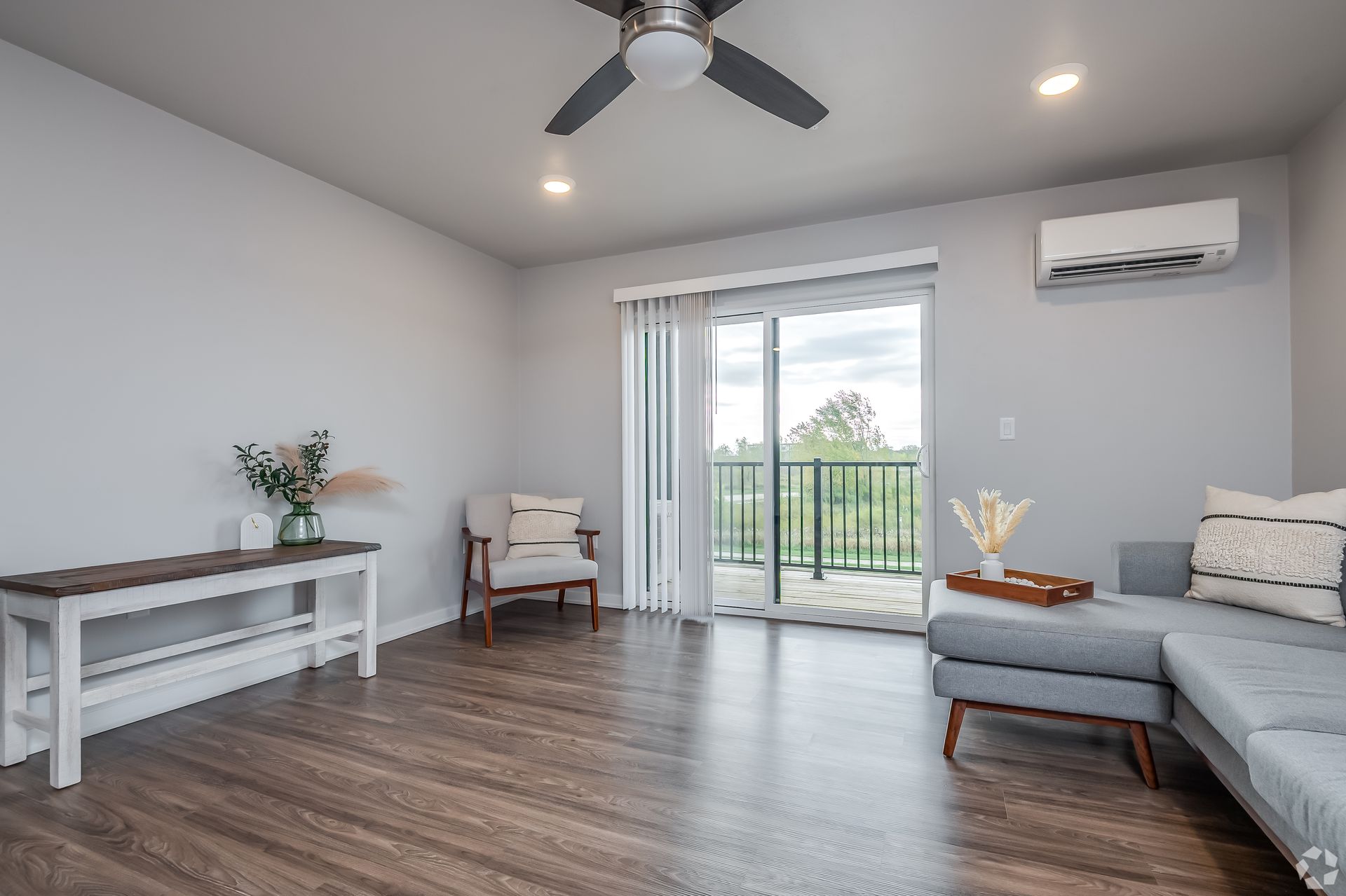 Bright living room with gray sectional, wood floor, sliding glass door, ceiling fan, and wall AC unit
