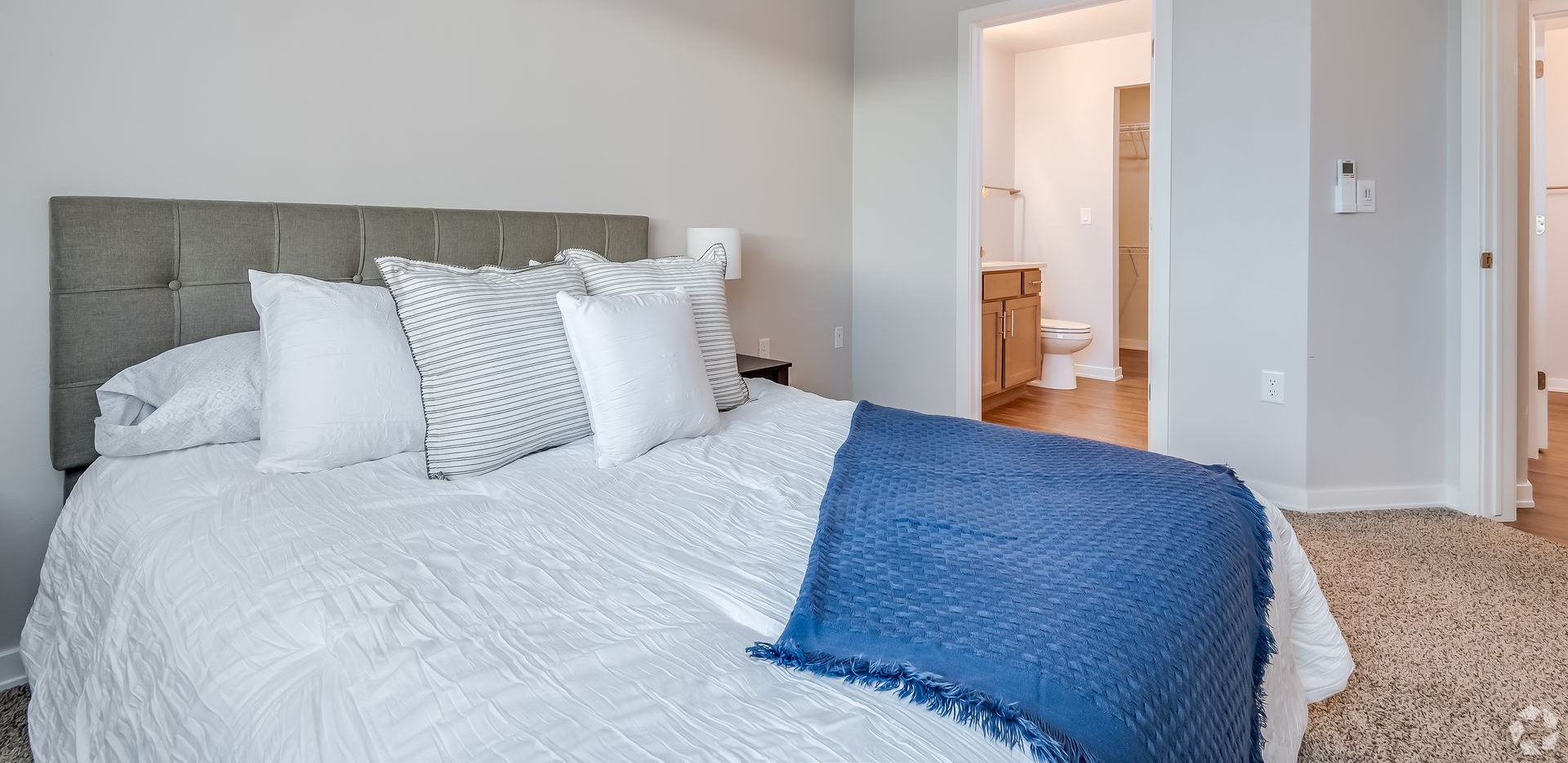 Bright bedroom with a neatly made bed, white bedding, blue throw, and open doorway to a bathroom.