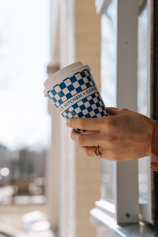 Hand holding a blue-and-white checkered takeaway coffee cup by a window
