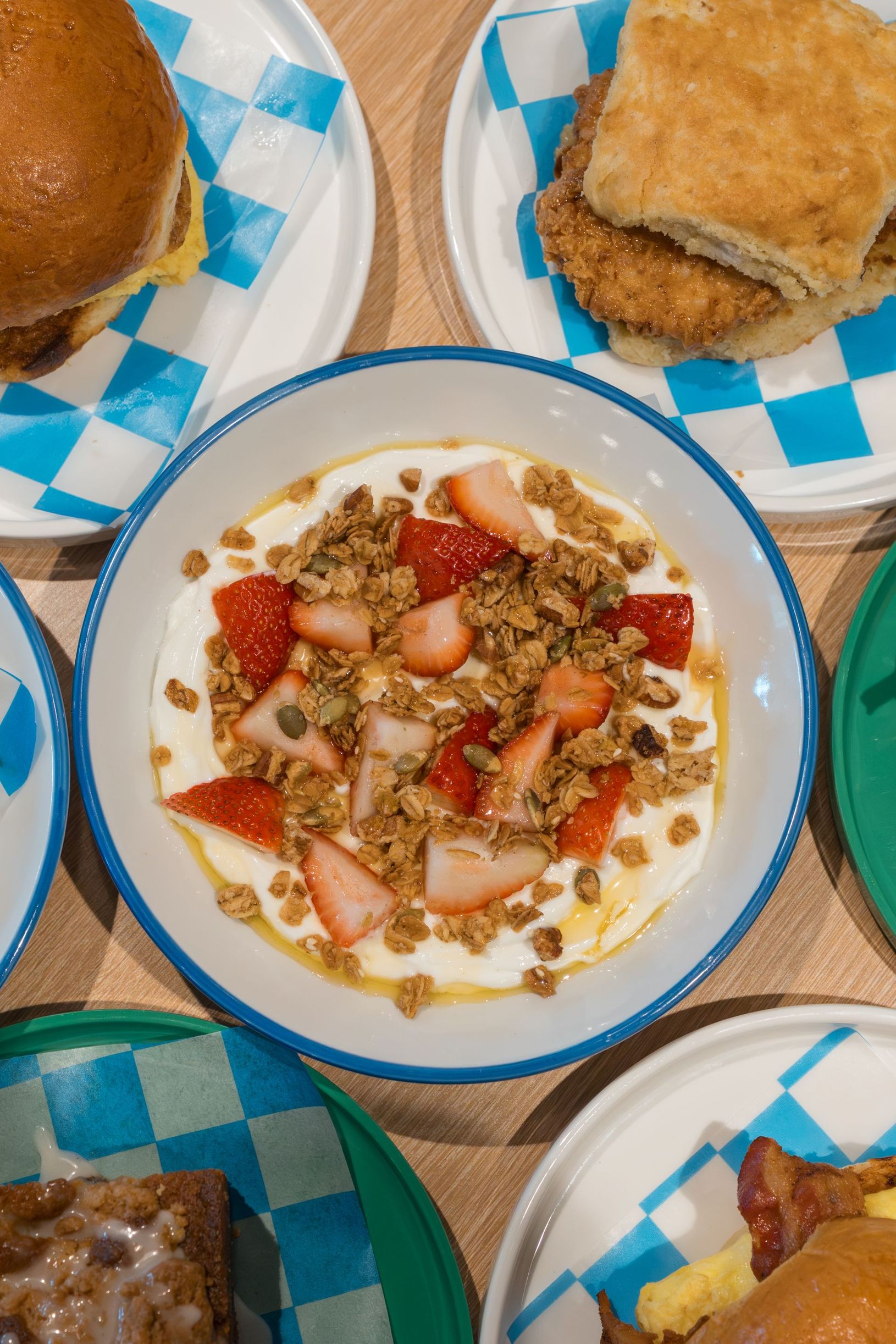 Top-down view of a meal spread with a central plate of food, bread rolls, and other dishes on a blue-checkered tablecloth